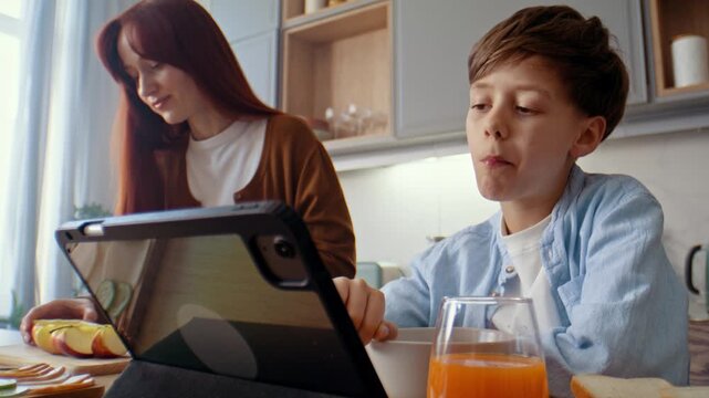 Boy eats corn flakes and watches content while mother cooks near