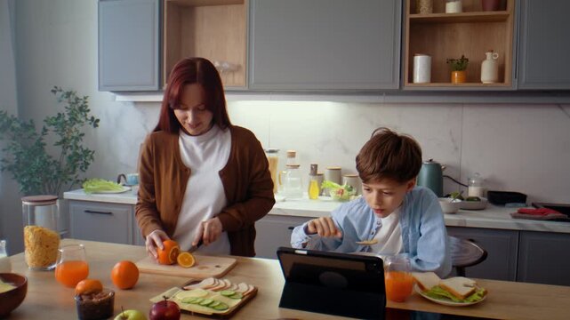 Woman cuts orange while son eats breakfast at digital tablet