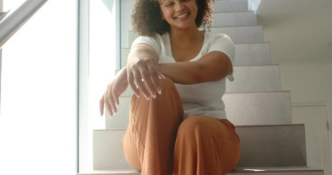 Sitting woman smiling and shifting weight on stairwell by window in white top, rust pants, bracelet