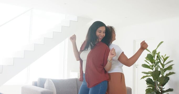 Diverse women friends smiling, syncing back-to-back moving at home by sofa rust white tops for fun