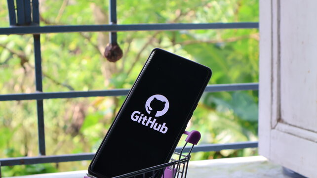 A smartphone with the GitHub logo on its screen is placed in a small shopping cart on a windowsill.