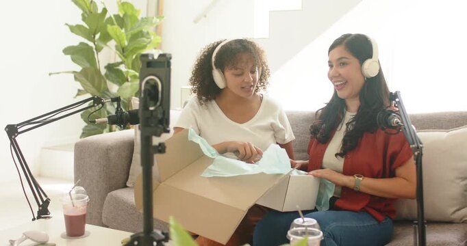 Diverse female hosts in studio opening box, revealing LED facial mask to smartphone and microphones