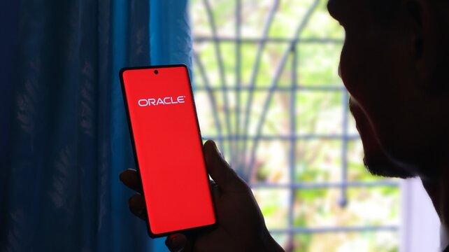 A person holding a smartphone with the Oracle logo on the screen in front of a window