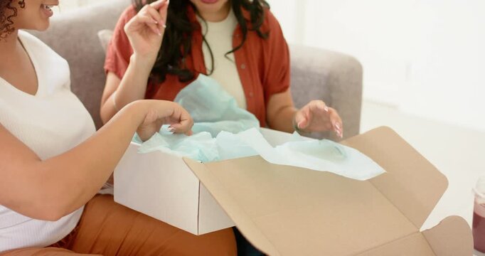 Diverse female friends on sofa opening box lifting tissue pulling LED mask and roller, checking fit