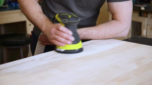 Carpenter using orbital sander on wooden plank. Close up of hands sanding wood in a workshop. Woodworking and craftsmanship concept