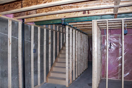Interior view of an unfinished basement construction site featuring wooden framing, exposed walls, and a staircase leading to the upper level