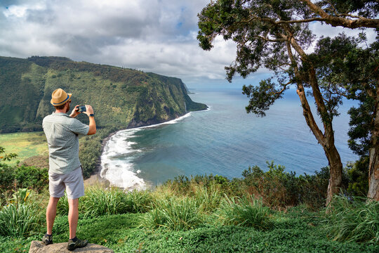 A male tourist standing and taking a photo of the Pololū Valley Lookout on the northern tip of the Big Island.
