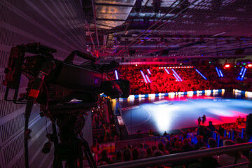 Professional broadcast camera on a tripod set up in a large sports arena, focused on a dimly lit handball court with spectators. © Dziurek