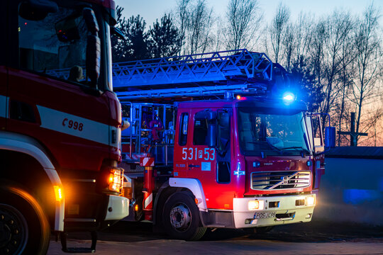 Two fire trucks, including a Volvo with a mechanical ladder, during a rescue operation at dusk. Visible blue emergency lights, operational numbers, and vehicle details against a sunset background.
