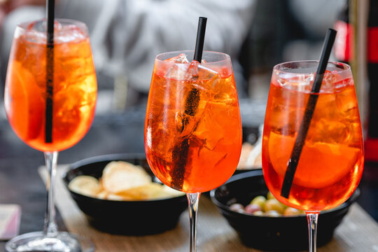 Vibrant orange Spritz cocktails with ice and citrus slices served in wine glasses with straws, accompanied by snacks in a casual dining setting