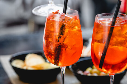 Vibrant orange Spritz cocktails with ice and citrus slices served in wine glasses with straws, accompanied by snacks in a casual dining setting