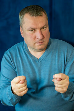 a middle-aged man with short hair making the "fig" hand gesture (dulya) with both hands while maintaining a serious expression against a deep blue textured background