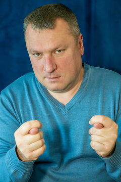 a middle-aged man with short hair making the "fig" hand gesture (dulya) with both hands while maintaining a serious expression against a deep blue textured background