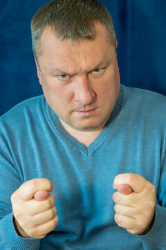 a middle-aged man with short hair making the "fig" hand gesture (dulya) with both hands while maintaining a serious expression against a deep blue textured background