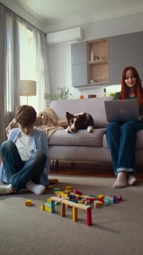 Boy builds wooden block bridge while woman works near lying dog
