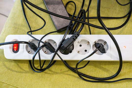 a messy tangle of black power cables plugged into a white european power strip with an illuminated red switch, resting on a green fabric surface in a home office.