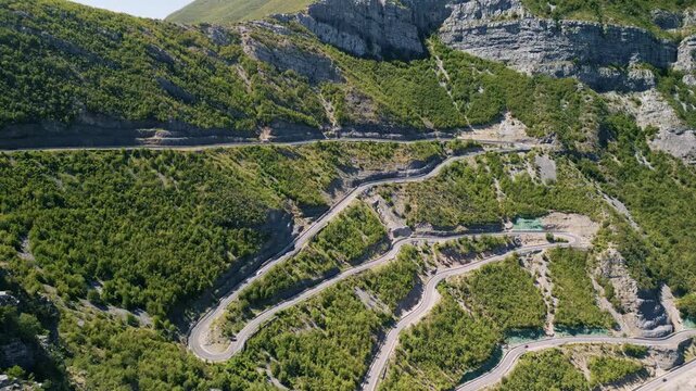 Aerial view of the incredibly scenic and winding SH20 mountain road in Albania with multiple switchbacks rugged cliffs and green slopes looking right over the traffic-filled highway during a sunny day