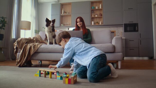 Boy builds colorful blocks while woman works near watching dog