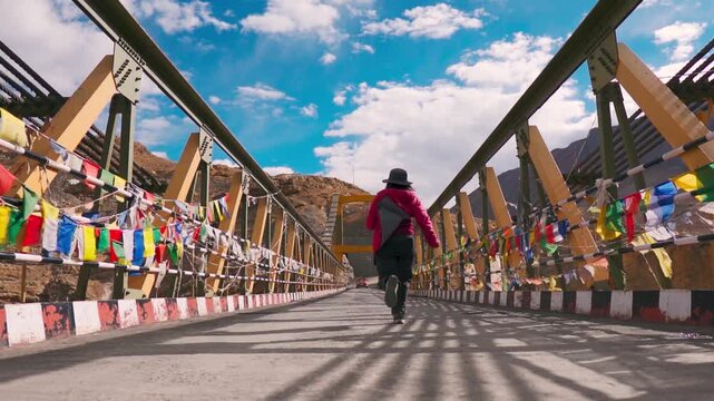 Indian woman running on Chicham bridge during sunny summer day in Spiti Valley, Himachal Pradesh, India. Buddhist prayer flags on suspension bridge in Spiti Valley. Travel and holidays background.