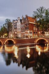 Obraz premium Illuminated Arched Bridge and Historic Dutch Houses Reflecting in Amsterdam Canal at Twilight