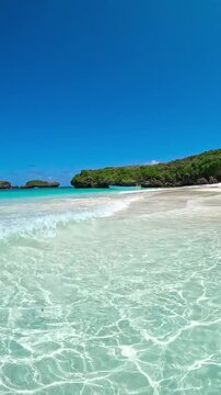 Turquoise ocean water ripples towards a white sand beach with adult men and women near green rocky islands under a clear blue sky in Watamu, Kenya.