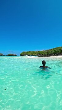 A young man swimming in turquoise water towards a white sand beach and green islands at low angle in Watamu, Kenya.