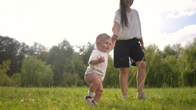 A mom helps her baby with the first lifestyle steps. Toddler child step. concept. A baby is taking the first steps in a. park. Mom helping her baby with his first steps in the park.