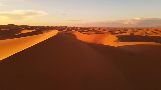 Wide angle shot of a vast desert landscape featuring undulating orange sand dunes bathed in warm sunset light with wind blowing sand off a high ridge under a clear sky.