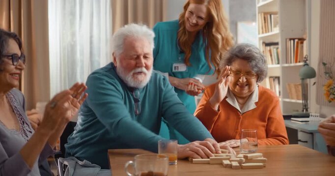 Seniors laughing as block tower collapses during game in assisted living home. Fun, spontaneous moment with joy, laughter and friendly social interaction indoors.