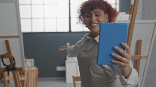 Woman holding blue tablet and gesturing with hand toward canvas in art studio while speaking and demonstrating brushwork to camera; remote teaching inspiration.