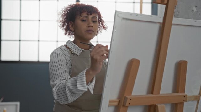 Woman painting canvas with brush at easel in studio, wearing apron and striped shirt; concentration creativity calm.