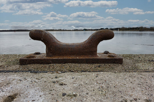 Ein rostiger Poller steht ruhig am Wasserufer unter wolkigem Himmel.