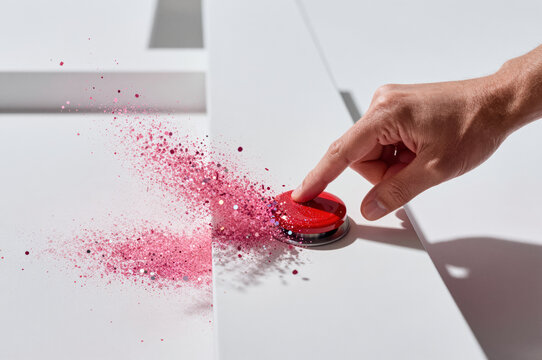 Human finger presses glossy red button on white surface, releasing pink glitter particles and metallic flakes in radial burst pattern under high contrast shadowing.