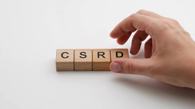 Hand arranging wooden blocks to spell CSRD on white surface. A symbolic presentation of corporate reporting and sustainability. Environmental compliance, organizational strategy, business regulations.