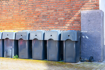 Obraz premium Row of dark gray plastic trash bins lined up against a weathered brick wall