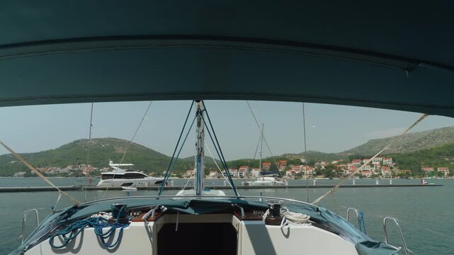 Boat Cockpit View Approaching Coastal Harbor, Under Bimini With Coiled Ropes On Bow, Calm Teal Water Reflecting Sunlight, Distant Mountains And Moored Sailboats Create