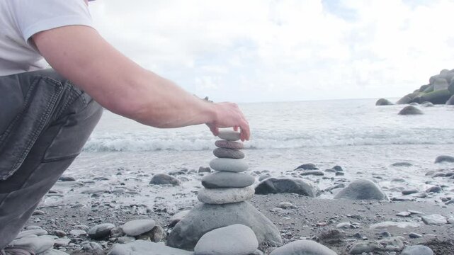 male hand carefully stacks Smooth zen-like pebbles, balanced pyramid against ocean waves, sea therapy on beach, Meditation and mindfulness, harmony, travel, and serenity coastal retreats