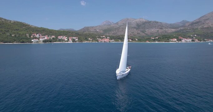 Sailboat Cruising By Waterfront Resort And Rolling Hills With Passengers Lounging On Deck, Bright Sun, Clear Water And Relaxed Holiday Atmosphere Captured By Drone
