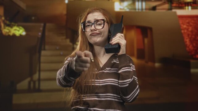 Redhead woman wearing glasses and striped sweater holds vintage handset and points finger at camera in building lobby staircase area; playful confidence.