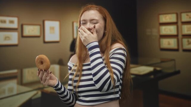 Redhead woman holding a donut, hand to mouth laughing in museum gallery amid framed prints and display cases; joy indulgence playfulness.