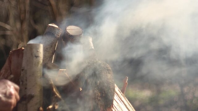 A close-up shows a man adding branches to a grill where the fire is starting to burn. The scene has natural spring light and a blurred forest background. Ideal for outdoor, camping, or BBQ content.