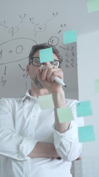 Thoughtful businessman wearing glasses concentrating on complex project plan. Analyzing strategy. Working with sticky notes and flowcharts on a clear glass board
