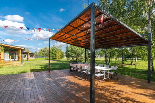 A group of white chairs and tables sits under a metal roof in a grassy area. Colorful flags hang above. Nearby are wooden buildings and green trees. The sky is clear and bright