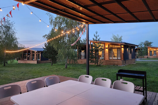 A group of cabins stands in a natural setting during the evening. String lights hang above a table with chairs. The atmosphere shows a place for social events and relaxation among trees