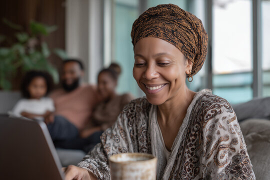 Smiling woman in headscarf using laptop at home with family in background; lifestyle portrait with warm natural light, concept of remote work, connection and wellbeing, ideal for ads and social media