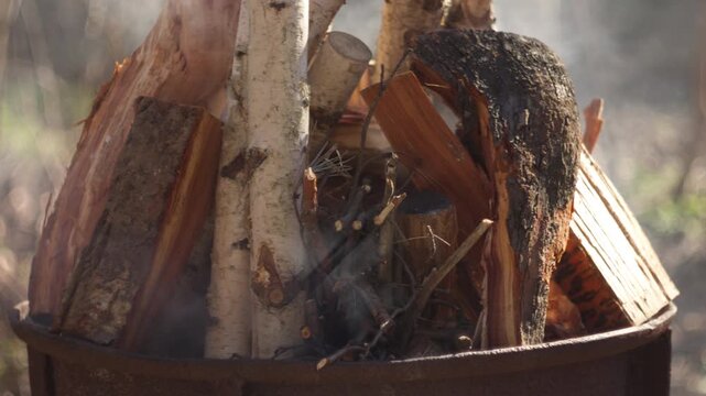Close-up of a man adding branches to a grill fire outdoors. Flames begin to burn with visible smoke and glowing embers. Natural spring light and a blurred forest background create a realistic camping 