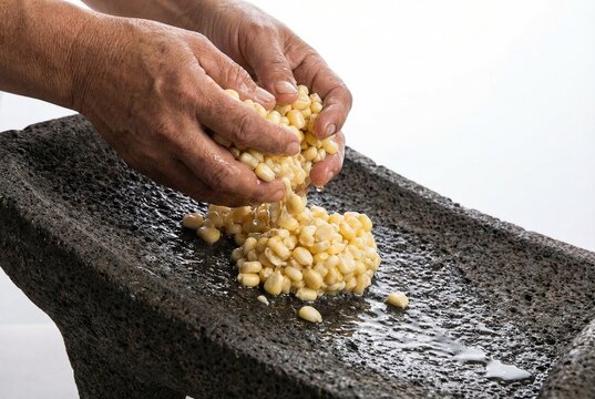 Hands squeezing water from fresh yellow corn kernels over a traditional volcanic stone metate