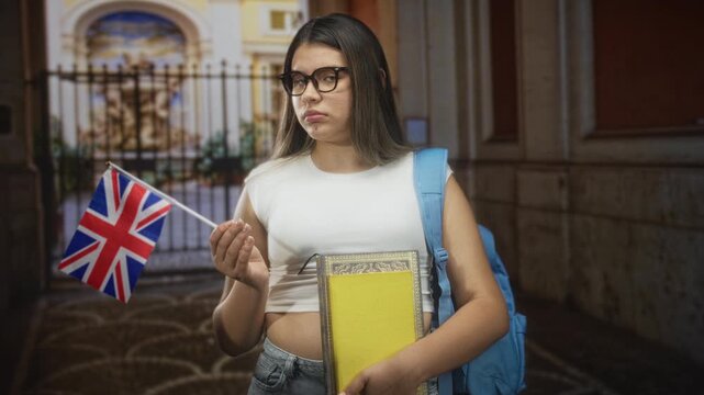 Young woman holding books and a uk flag, looking at the flag with a pensive hand gesture in a building entrance; homesickness study abroad.