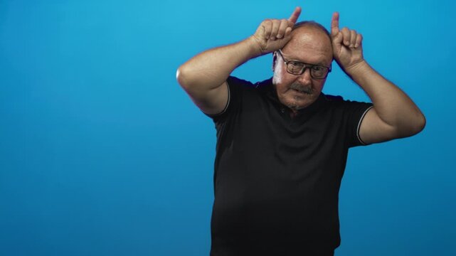Man senior hispanic wearing black polo and glasses points index fingers to head like horns in blue studio; playful mischief.