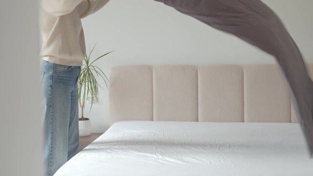 Woman shakes gray duvet over bed in bedroom interior